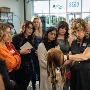 Female hair expert demo on a wig model surrounded with hairstylists